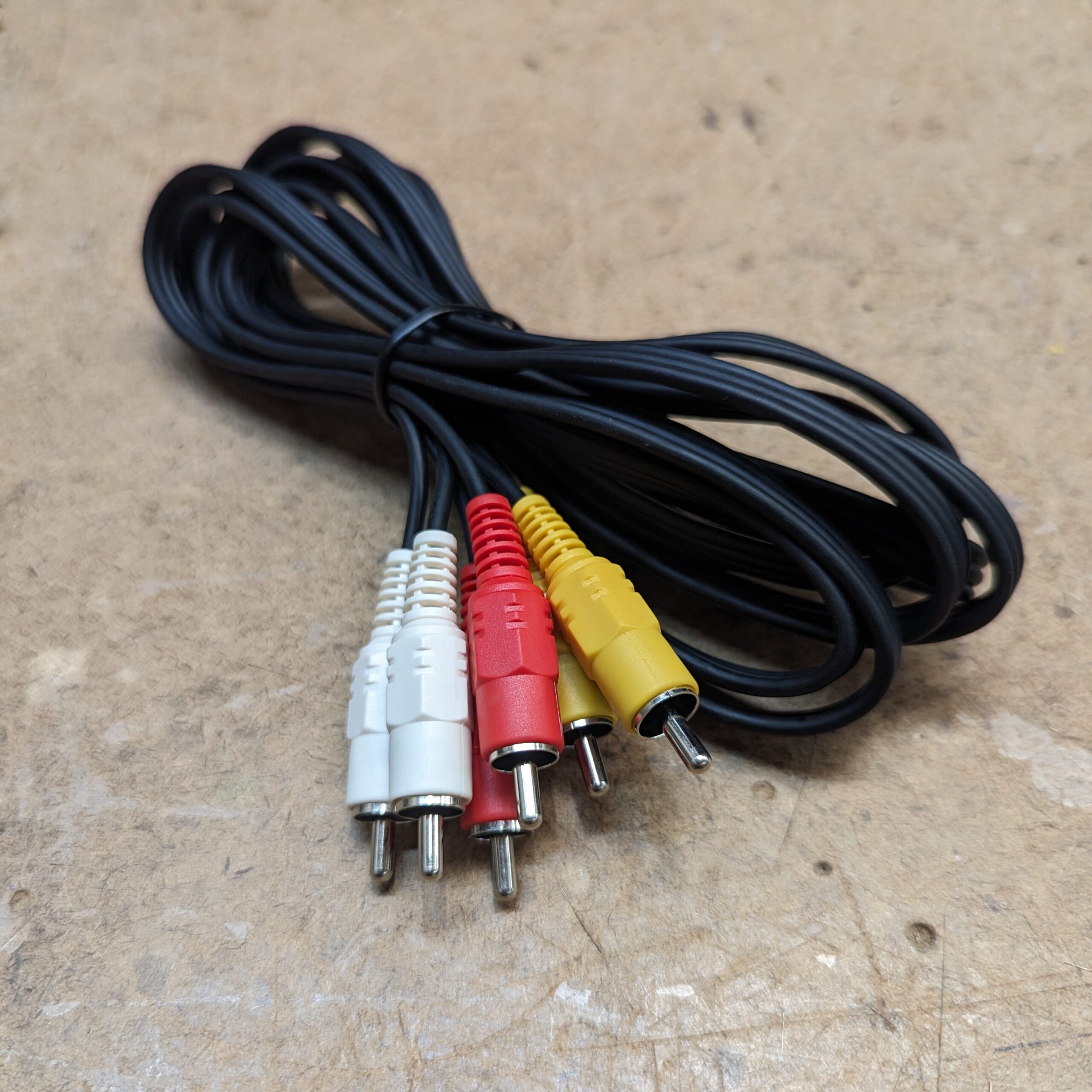 A coiled black composite video cable with three colored RCA connectors at one end – white, red, and yellow – lies on a textured, light brown surface.