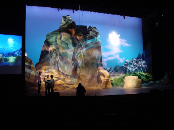 The stage is brightly lit, focusing on a rocky formation in front of a blue sky background. Three figures are standing on the left side of the stage, while another sits on the floor on the right side. A smaller video screen is visible to the left, showing the same scenery as the background.