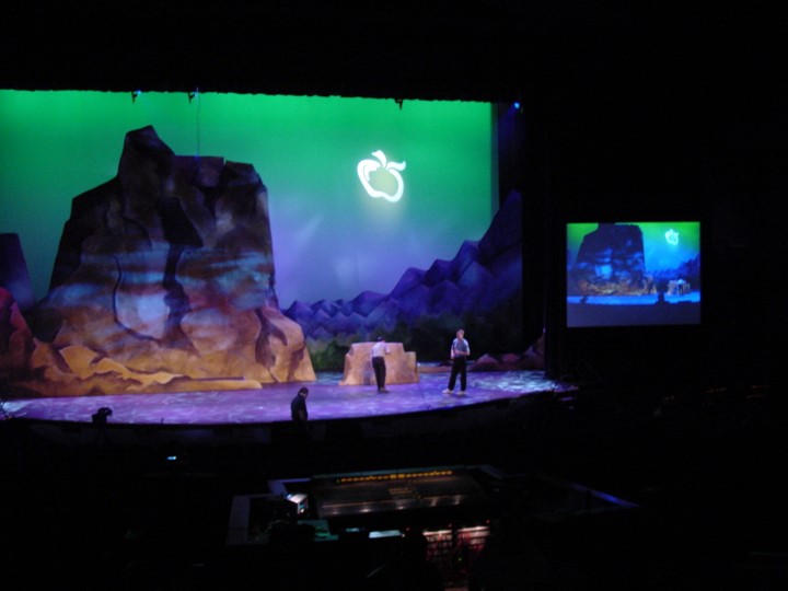 A wide shot of a theatre stage in a dark room. The stage is lit with a green backdrop showing rocky terrain and a glowing apple logo in the center. There are three people on the stage, one to the right and two at center stage. A smaller video screen to the right shows the same background. The audio and lighting control board is in the foreground.