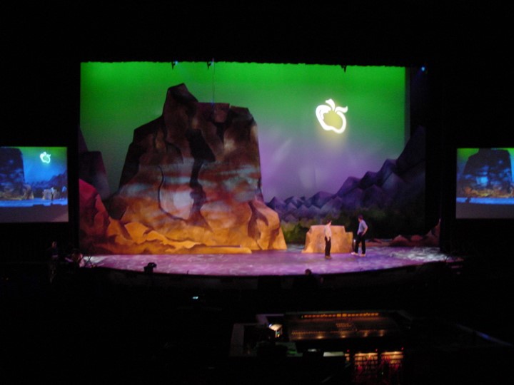 Similar to the first image, a wide shot of the stage with a green background, glowing apple logo, and rocky terrain. The lighting is focused in the center stage, where two people are standing. Two smaller screens appear on the left and right side displaying the same image of the rocky terrain. The audio and lighting control board is in the foreground.