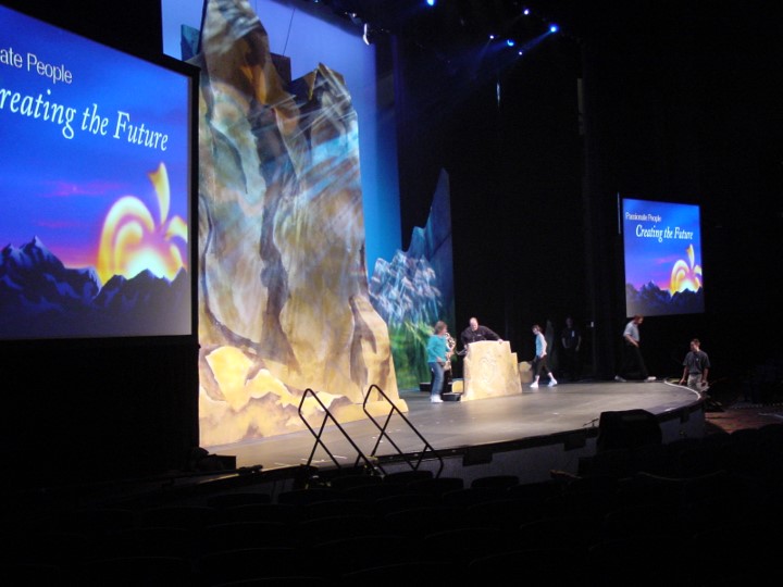 A stage with rocky props and a backdrop showing a mountainous background. A few people are on stage, with some standing behind a rocky formation. On the right and left side are two smaller screens displaying “Passionate People Creating the Future”.