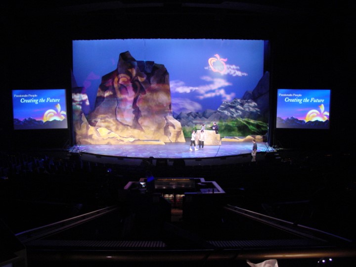 A long shot of the stage, showing the same blue-toned mountain scenery with a sun and cloud. Two screens now appear on each side of the stage displaying “Passionate People Creating the Future”. The audio and lighting control area is visible in the foreground, and some small figures are visible on stage.