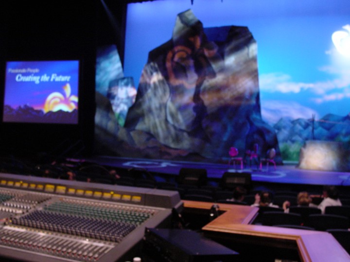 A closer view of the same theatre stage, the lighting has changed to a blue-toned background, now with a detailed sky and sun behind the rocky formations. A smaller screen on the left displays “Passionate People Creating the Future” with an apple graphic. In the foreground, the soundboard is in the focus with rows of seats in the background.