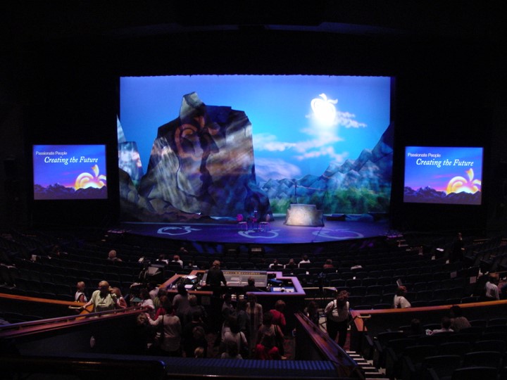 A wide shot of the stage and audience. The stage is lit with a blue-toned background and a detailed sky with a sun. Two smaller screens display the words “Passionate People Creating the Future” on each side of the stage. The audience is starting to fill in the rows of seats.