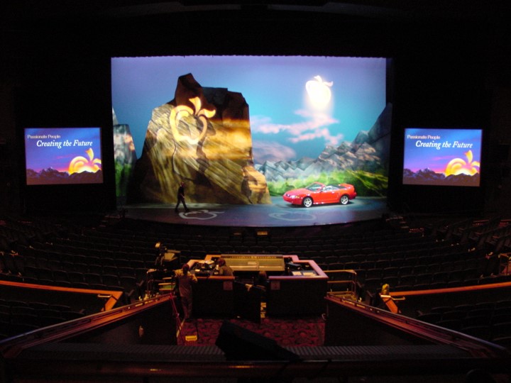 A shot from the back of a theater. The stage displays rocky scenery and a blue sky, with an animated sun and cloud. A red car is parked on the left side of the stage. Two side screens display “Passionate People Creating the Future”, with a stylized flower. The audio and lighting control area is visible in the foreground.