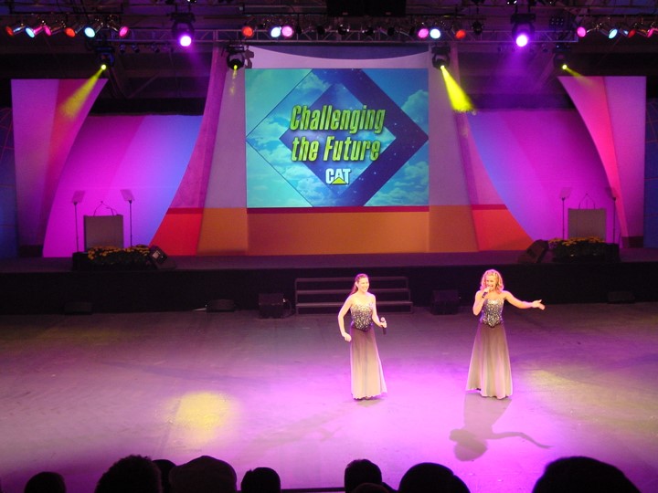 A wide shot of two women singers center stage, part of the entertainment for a Caterpillar dealer event. The stage is brightly lit with pink and purple hues, and a large screen displays the CAT logo and "Challenging the Future