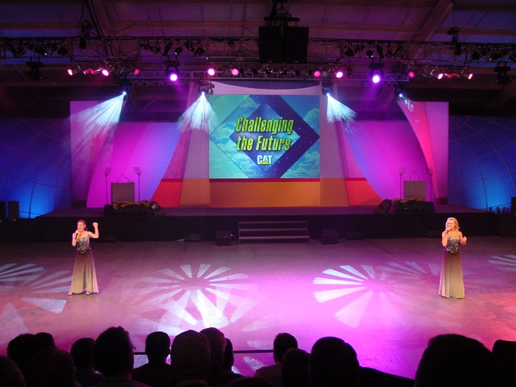 Two singers perform on a brightly lit stage for a Caterpillar dealer meeting. A large screen displays the CAT logo and "Challenging the Future".