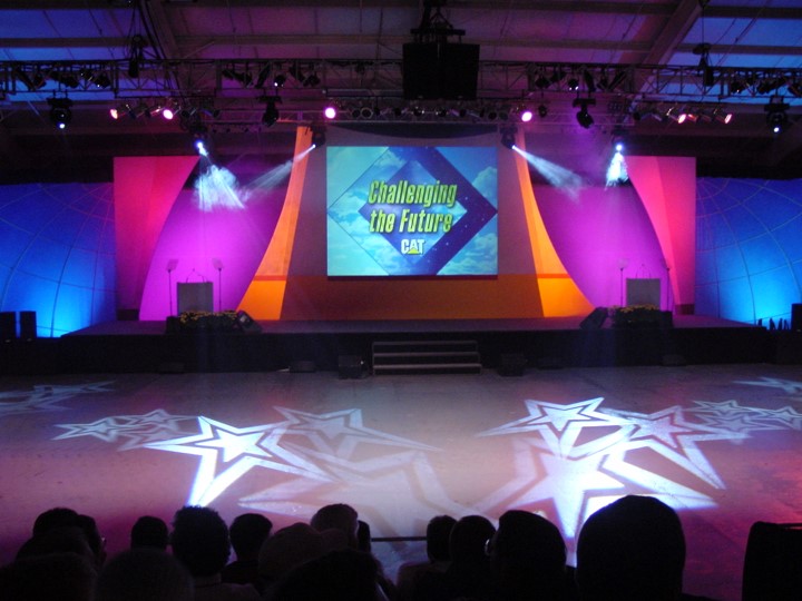 The event stage for a Caterpillar dealer meeting is brightly lit with pink, purple, and yellow hues. Star-shaped light patterns are projected onto the floor and a screen displays the CAT logo and the slogan "Challenging the Future