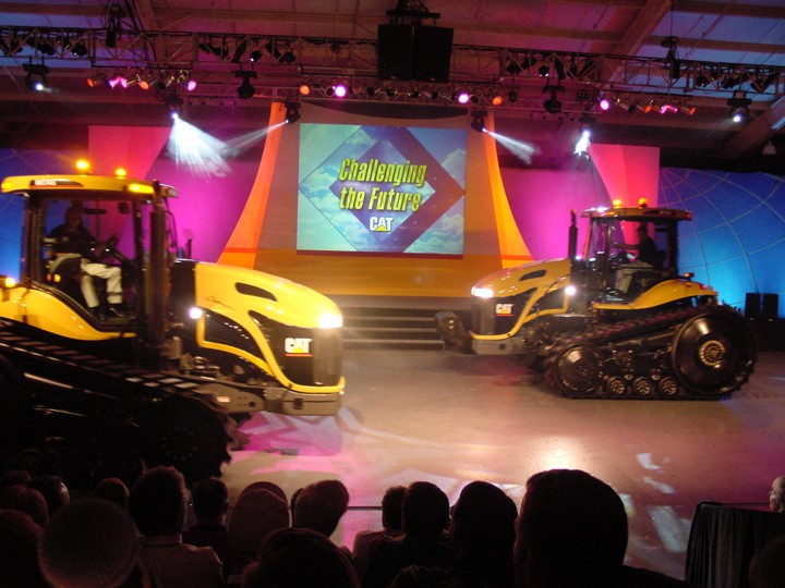 Two Caterpillar 700 series tractors are revealed on the stage of a dealer meeting, accompanied by dramatic lighting and a large screen displaying the "Challenging the Future" message.