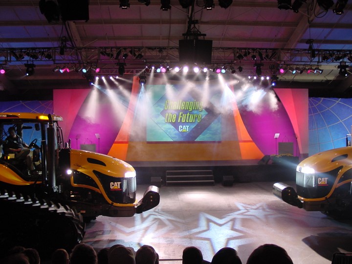 Two Caterpillar 700 series tractors are featured on a stage with dramatic lighting during a dealer event. A large screen behind them reads "Challenging the Future CAT". Star-shaped light patterns are also visible on the stage floor.