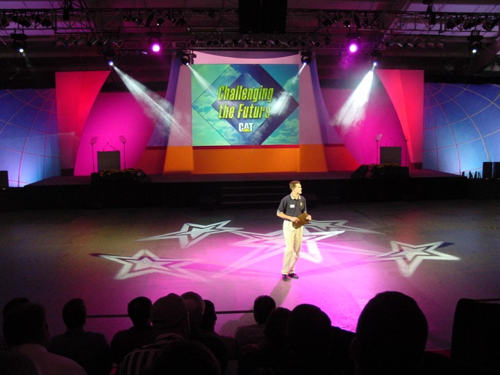 A man stands on the stage, spotlighted against a backdrop of star-shaped projections on the floor, during a Caterpillar dealer meeting, where a screen reads "Challenging the Future CAT".