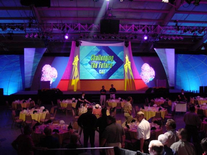 A long view of the event stage during a Caterpillar dealer meeting, taken from a location with tables for dining. People stand at and sit around the tables as the stage with the "Challenging the Future" screen is prepared for the next part of the event.