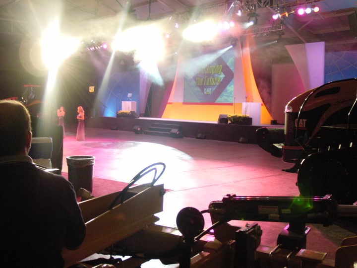 The event space for a Caterpillar dealer meeting seen from the side of the stage. The scene includes two singers performing on a brightly lit stage as well as some of the yellow Caterpillar equipment used in the "tractor ballet."