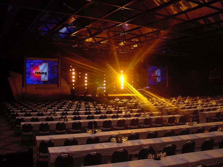 A photo of the Discover Card meeting venue at the Contemporary Hotel in Orlando from the rear of the audience, illuminated by a bright yellow stage light with rays shooting out. There are two large screens on each side showing the number "1" and the word "POWER". Rows of tables with white tablecloths fill the room in front of the stage.