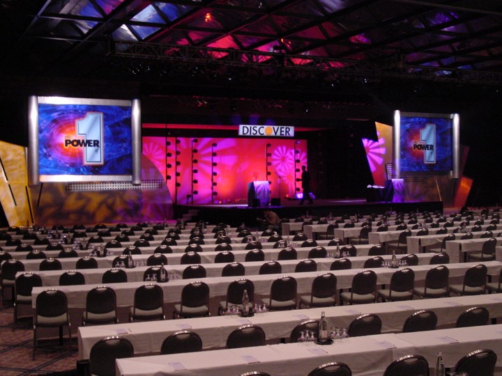 An overview of the Discover Card meeting venue at the Contemporary Hotel in Orlando. Rows of white-clothed tables and chairs face a stage with a "DISCOVER" banner. The stage has two side screens showing the number "1" and the word "POWER". Lighting is bright on stage.