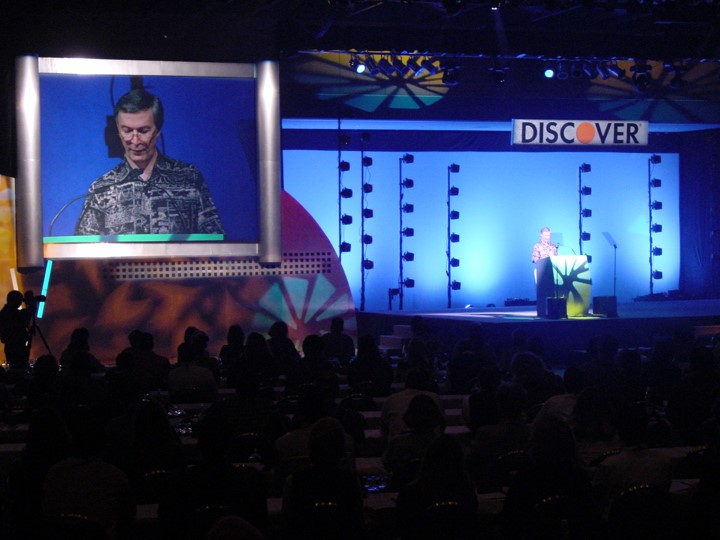 A man is speaking at a lectern on stage during the Discover Card meeting in Orlando. A large screen displays a close-up image of the speaker. The stage backdrop is brightly lit with blue light. The rows of the audience are visible.