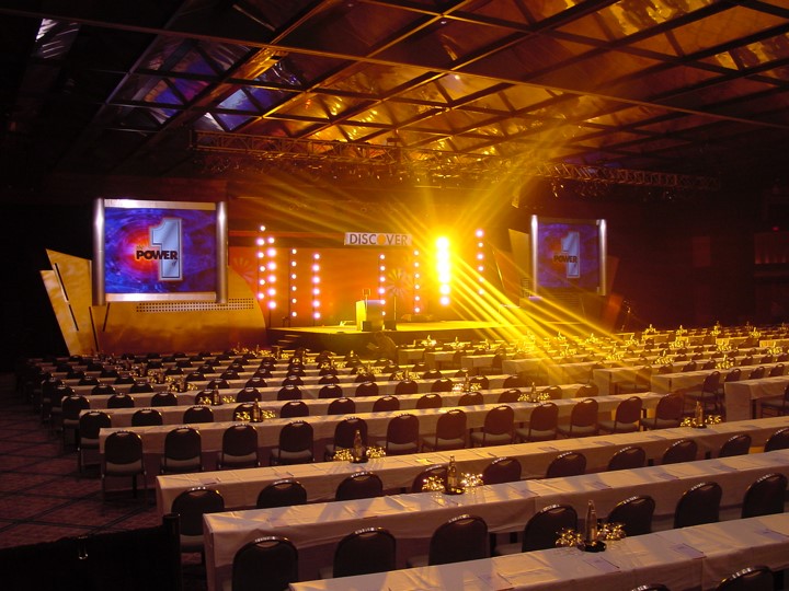 A photo of the Discover Card meeting venue at the Contemporary Hotel in Orlando, illuminated by a bright yellow stage light. Two large screens are on each side of the stage showing the number "1" and the word "POWER". The room has rows of tables with white tablecloths and chairs for the audience.