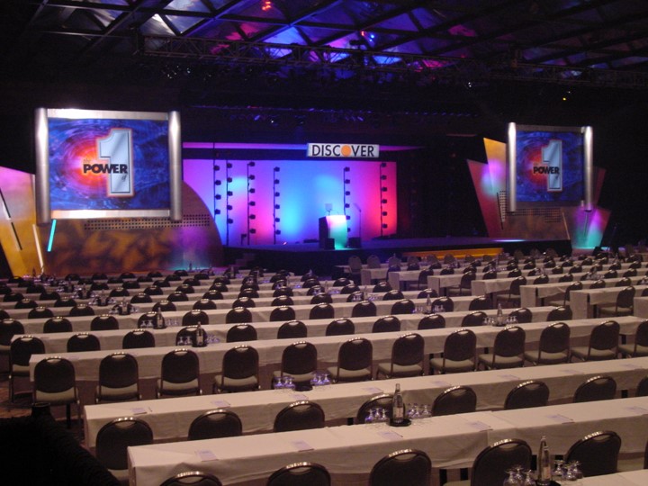 An overview of the Discover Card meeting venue at the Contemporary Hotel in Orlando. Rows of white-clothed tables and chairs fill the room in front of the stage. The stage has a "DISCOVER" banner, and side screens showing the number "1" and the word "POWER" are displayed. The backdrop is illuminated with colorful pink and blue lights.