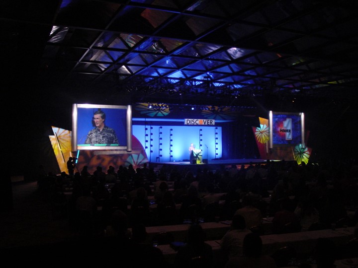 A man is on stage speaking at the Discover Card meeting. A large screen on the left side of the stage shows a close-up of the speaker. Behind the man on stage, is a light blue background with the "DISCOVER" banner over him. The rows of tables fill the room before the stage.