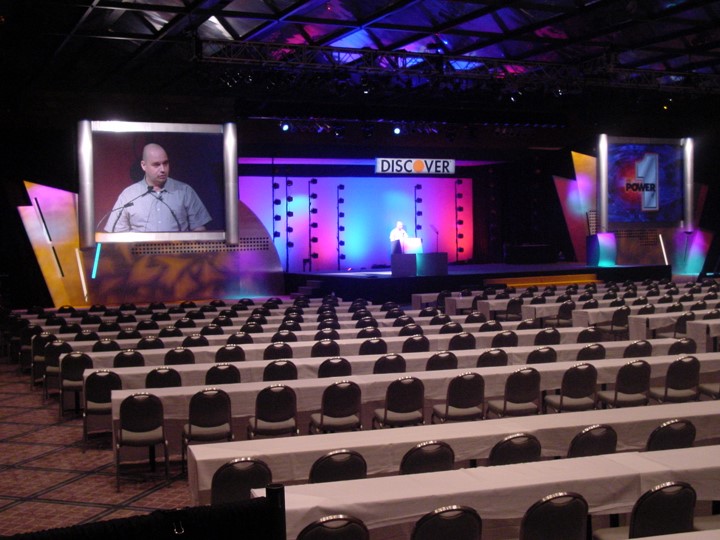 A man is speaking on the stage at the Discover Card meeting. A large screen displays a close-up image of the speaker. There is a "DISCOVER" banner on stage, which is lit with vibrant pink and blue lights. Rows of white clothed tables fill the room in front of the stage.