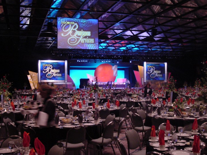 A Discover Business Services conference room at the Contemporary Hotel in Orlando is set for a dinner with red napkins and black tablecloths. The stage has the words "DISCOVER BUSINESS SERVICES" on the screens and is set with flower center pieces.