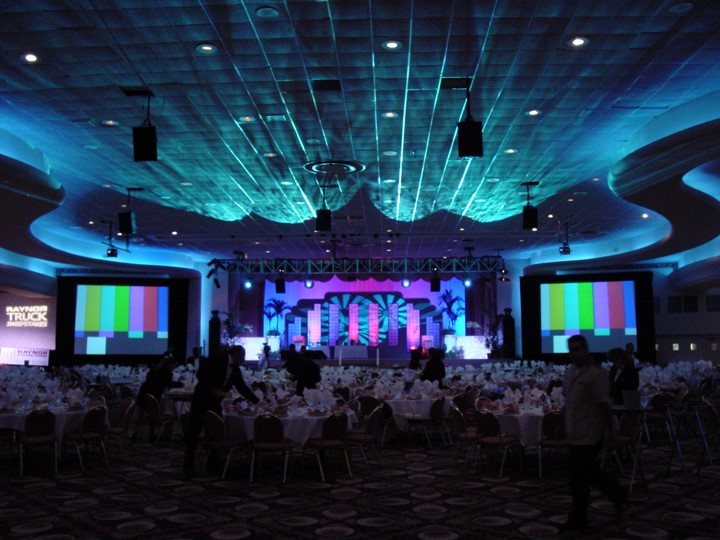 View of the Raynor Garage Door meeting setup in the Fontainebleau Resort, Miami Beach. The image shows a large room with round dinner tables covered in white tablecloths and set for a meal, and two side screens displaying test patterns on the stage. The ceiling is lit with turquoise and the stage features decorative panels lit with pink, purple and blue. The meeting room appears to be under preparations for the dinner portion of the event