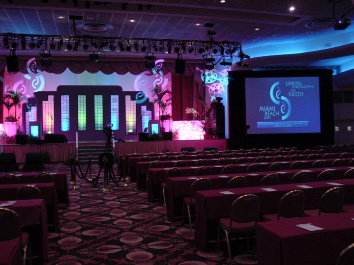 Wide shot of a large conference room at the Fontainebleau Resort in Miami Beach, set up for a Raynor Garage Door meeting. A stage is adorned with set pieces, pink curtains, and potted plants. A central projection screen displays the 'Linking Strengths for Success' conference logo, while rows of tables and chairs are arranged for the audience. Stage lighting, including blue, purple, and pink, illuminates the space.