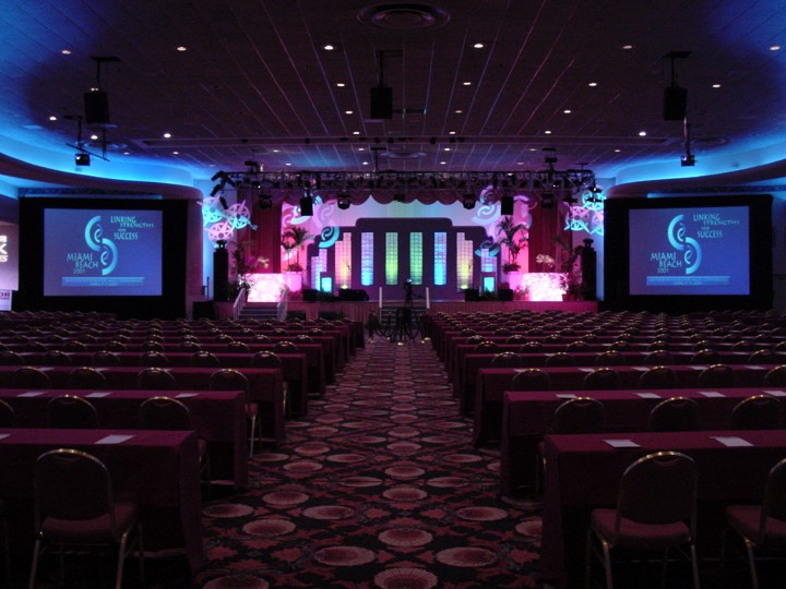 Wide angle view of a large conference room at the Fontainebleau Resort, Miami Beach, set for a Raynor Garage Door meeting. In the foreground there are rows of tables and chairs covered in maroon cloths. In the background there is a stage and two large off-stage screens projecting 'Linking Strengths for Success'. The stage is decorated with colorful lights, plants, and modern decorative elements.