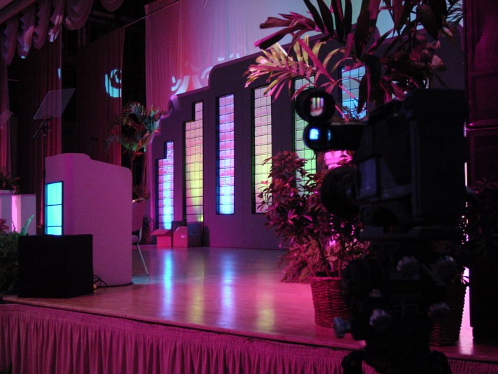 Close-up, low-angle view of the stage from behind a camera tripod at the Raynor Garage Door meeting at the Fontainebleau Resort, Miami Beach. The stage is adorned with modern decorative panels lit in colors including pink, blue, and purple. There is a small podium at the front left of the stage. Potted plants are placed at various locations.