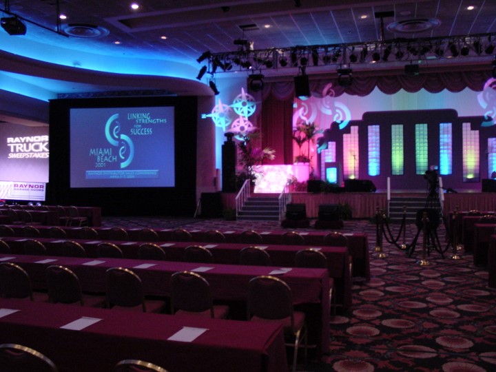 Image of a meeting space in the Fontainebleau Resort, Miami Beach, prepared for the Raynor Garage Door meeting. There is a stage with various set pieces and lighting with the text 'Linking Strengths for Success, Miami Beach, 2001' projected onto a large off-stage screen. Rows of tables covered with maroon cloths and chairs for conference attendees are set up in the foreground. There are also some camera tripods in the scene.