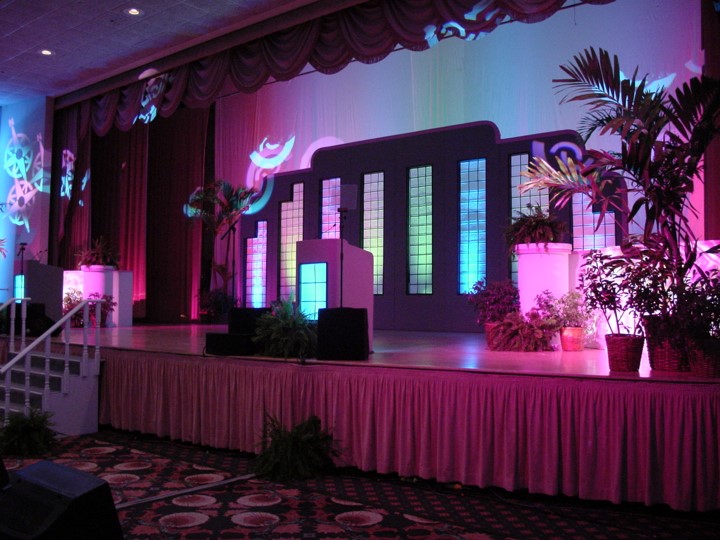 Close-up view of the stage at the Raynor Garage Door meeting held at the Fontainebleau Resort in Miami Beach. The stage features a set decorated with a pink curtain backdrop and modern, colorful cut-out shapes. A lit podium stands at stage left. Potted plants are positioned on and around the stage, all lit with pink and blue lighting.