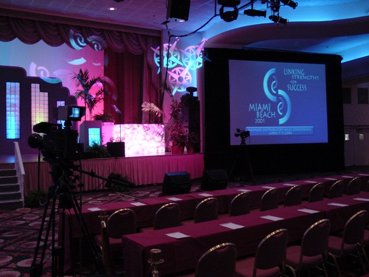 View from the front of the conference room of the stage set-up and a screen projection at the Raynor Garage Door meeting at the Fontainebleau Resort, Miami Beach. A central screen displays the conference logo and a stage with a pink curtain backdrop and modern, colorful cut-out shapes. Camera equipment and rows of tables are set up for the event.