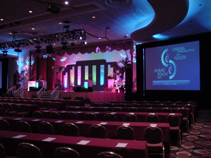 Image of the stage setup for the Raynor Garage Door meeting at the Fontainebleau Resort in Miami Beach. The view is looking at the stage from the front of the room, showing the screen, podium, main stage and audience tables covered in maroon cloths. The stage features pink curtains and a colorful modern cutout set with potted plants. Off-stage, on the right and left, are two large projection screens displaying the conference logo.