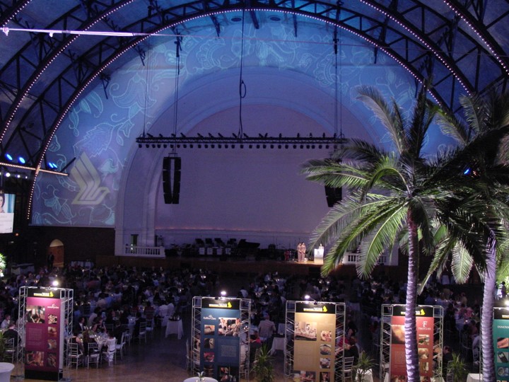 Interior of the Aon Ballroom at Navy Pier, set up for a Singapore Airlines event. A large stage is framed by a projected floral design and the Singapore Airlines logo on the wall. Four 20 ft palm trees flank the seating area, where guests sit at tables. Informational panels on stands are visible near the foreground. Lighting creates a blue-purple ambiance.