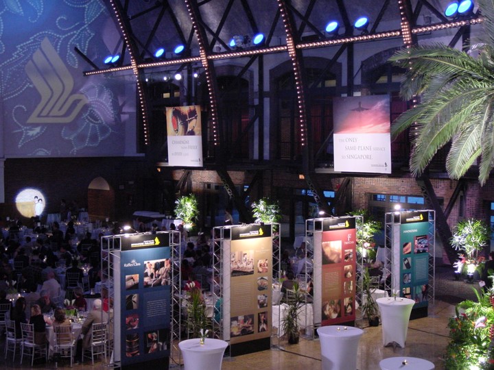 Wide view of the Aon Ballroom at Navy Pier set up for a Singapore Airlines event. Palm trees frame the scene. Display panels stand in the center, highlighting information about the new flight to Singapore. The ceiling is visible with beams and lighting. Banners in the windows feature the Singapore Airlines logo.