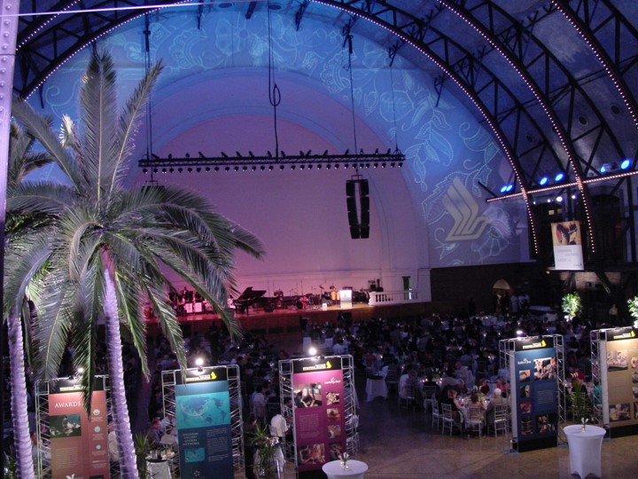 Elevated view of the Aon Ballroom at Navy Pier, showcasing a dinner event for Singapore Airlines. Palm trees flank the space. A projected floral pattern and the Singapore Airlines logo is on the arched ceiling and stage area behind where an orchestra plays. Informational panels line the central aisle of dining tables. The color wash is a light purple.