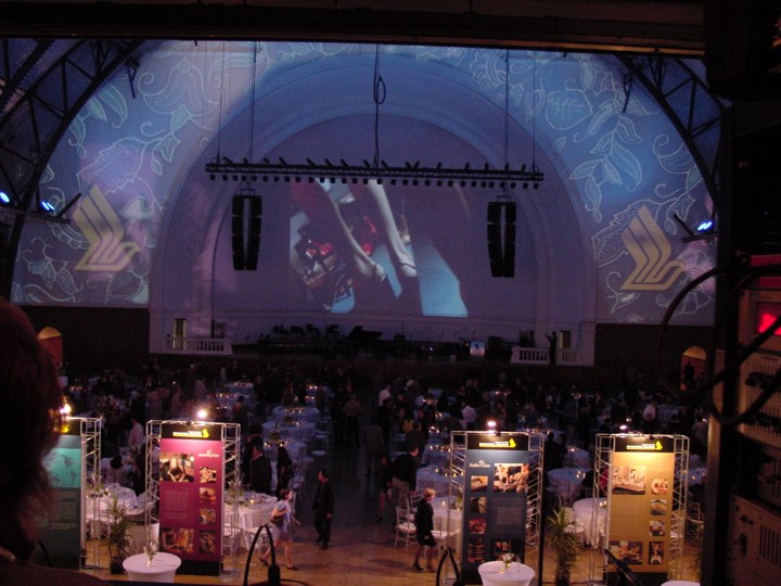 Wide shot inside the Aon Ballroom at Navy Pier, during the Singapore Airlines event. A large screen projection shows people in traditional dress. Guests are seated at round tables and an orchestra is visible on the stage. The ceiling is illuminated with a blue wash. A floral pattern is projected onto the arched ceiling and the Singapore Airlines logo.