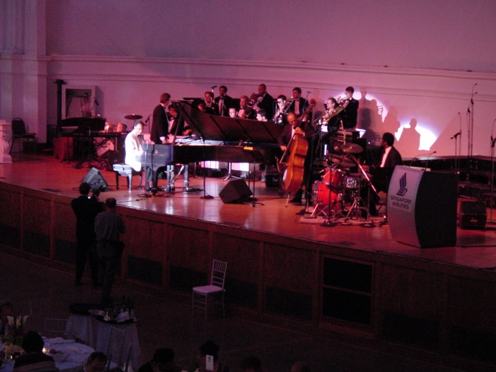 Close up of a band playing on a stage during a Singapore Airlines event. The band includes a grand piano, drums and several horns. The stage is bathed in red light, the Singapore Airlines logo on a podium.