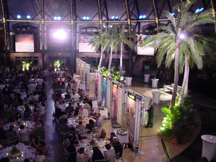 Full view of a Singapore Airlines event at the Aon Ballroom at Navy Pier. Guests are at dinner tables leading into the hall with palm trees and informational panels. The ceiling has lighting and beams and banners are on the walls. 