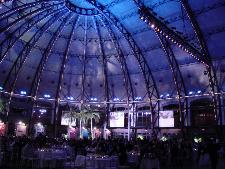 High, wide angle view of the ceiling in the Aon Ballroom at Navy Pier at the Singapore Airlines event, with the structural beams highlighted by blue lighting. The dining area, palm trees, and four information panels are visible below the vaulted ceiling. Guests sit at round tables.