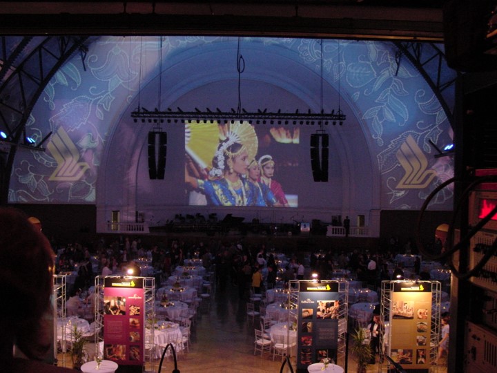 Elevated shot of the Aon Ballroom at Navy Pier during the Singapore Airlines event. A large screen shows dancers in traditional dress, and floral design and the Singapore Airlines logo projected onto the ceiling. The space is set with round dining tables, four display panels and an orchestra is on the stage.